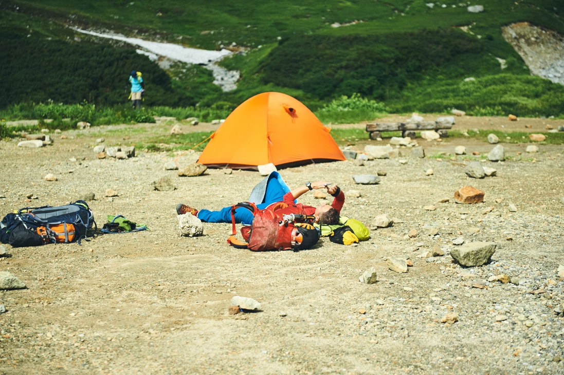 A hiker relaxes under an orange tent on a rocky plateau with green mountains and a river visible in the background.
