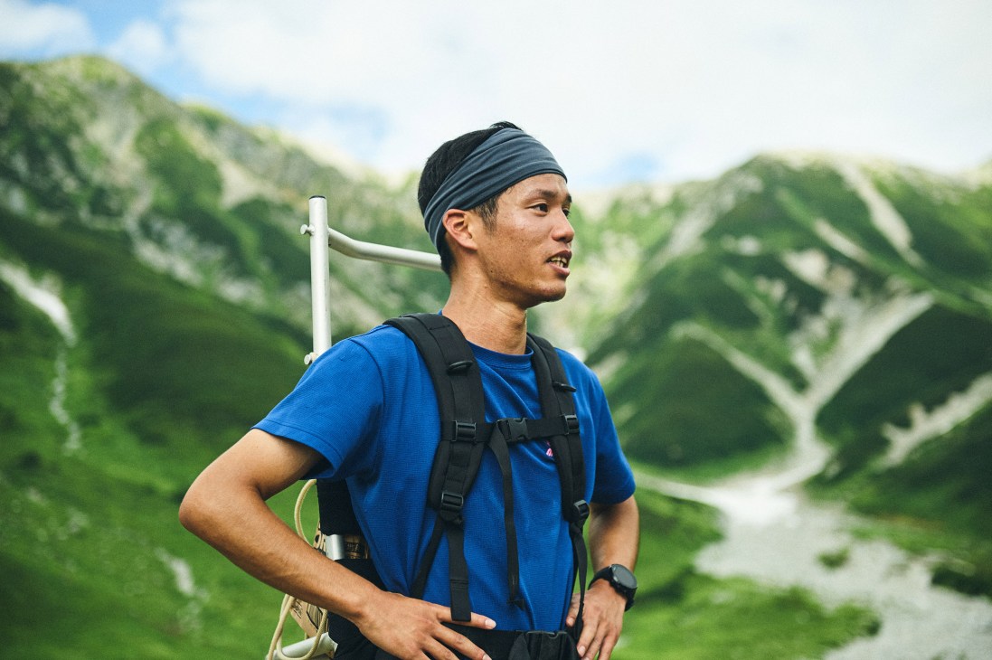 A hiker in a blue shirt and dark headband carries a white pipe while looking out over green mountains in the Tateyama range.