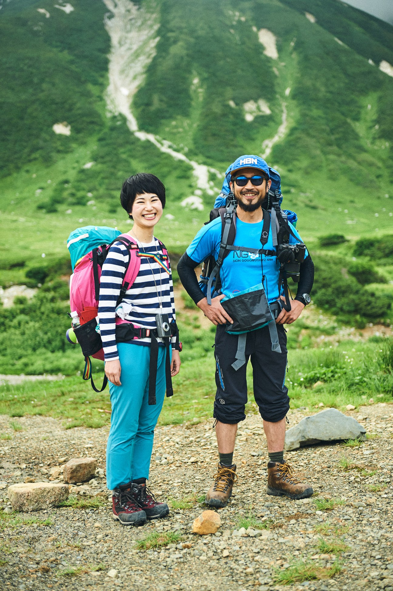 Two hikers in colorful sportswear stand on a mountain trail with verdant valleys behind them.