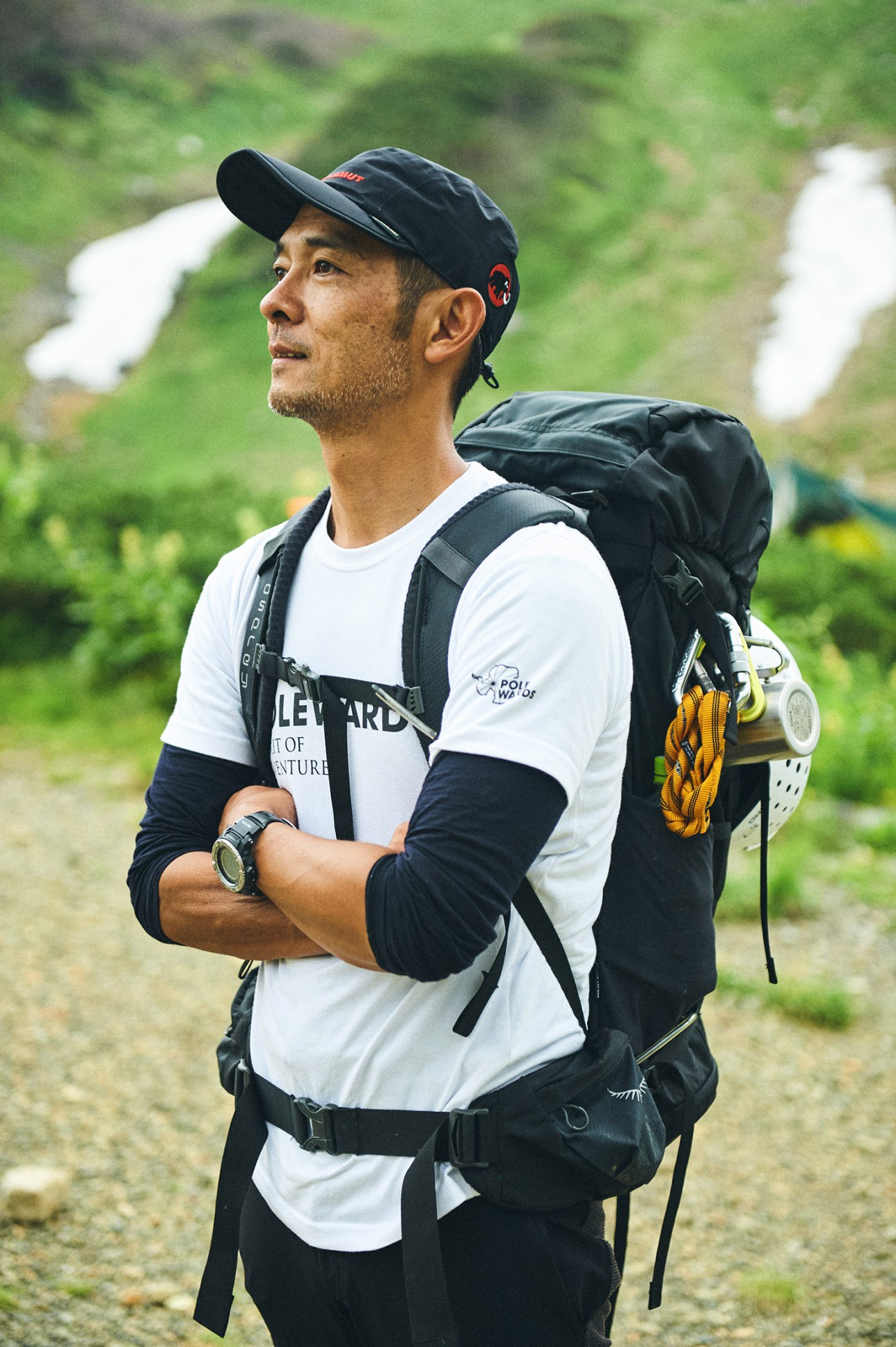 Man wearing mountain climbing gear including backpack, cap, and rope equipment standing on a trail with green hillside background.