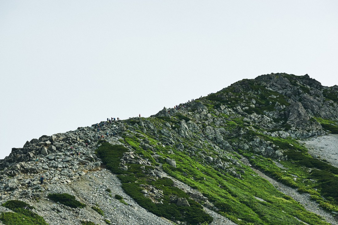 Hikers in single file line walking along a steep rocky ridge with green vegetation on the Tateyama mountain range.