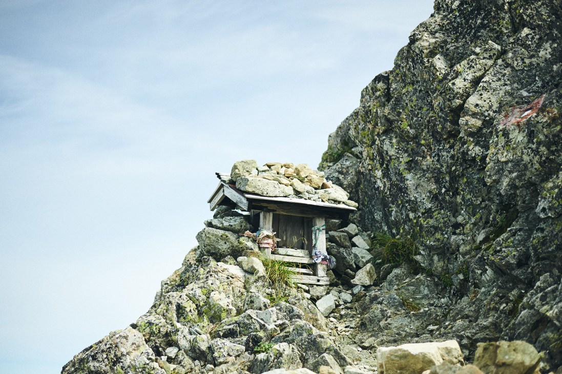 A small wooden shrine nestled among rocky peaks on a mountainside with moss-covered stone cliffs.