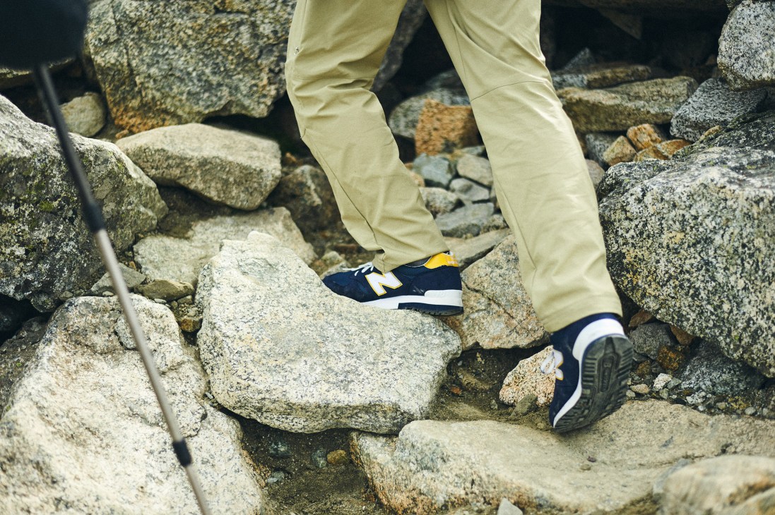 Close-up of hiker's legs wearing pale yellow trekking trousers and New Balance sneakers on rocky mountain terrain.