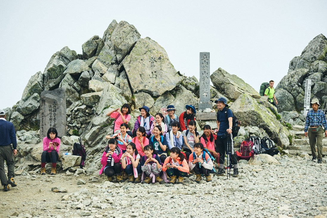 Group of hikers in colorful mountaineering outfits pose together on rocky summit of Mount Oyama.