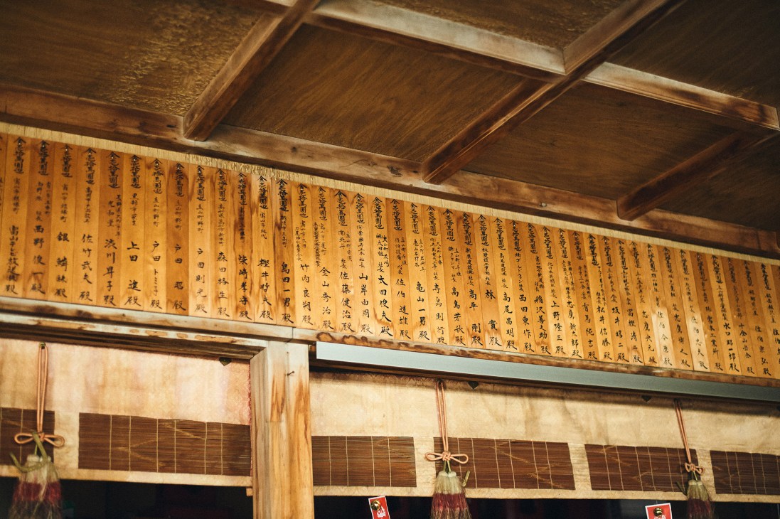 Wooden plaques with Japanese calligraphy hanging on interior shrine walls beneath wooden beams.