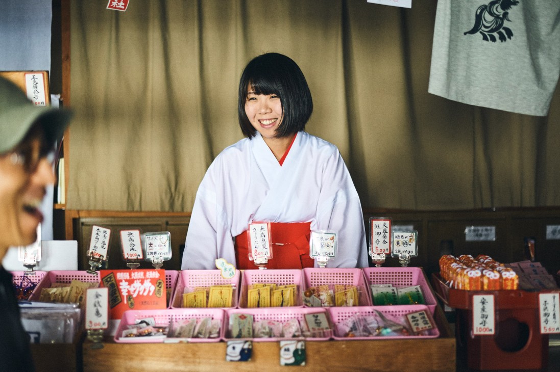Woman in white kimono with red obi smiling behind counter displaying packaged snacks and souvenirs.