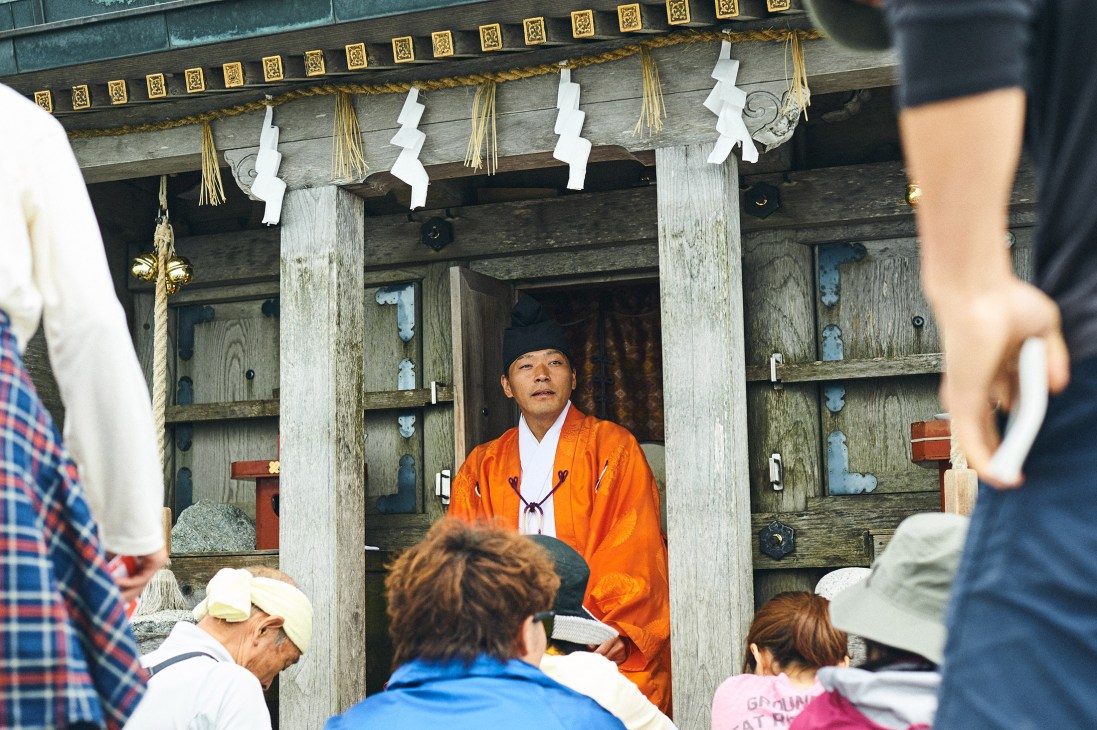 A Shinto priest in an orange robe performs a blessing ceremony inside a small wooden shrine for a group of pilgrims at high altitude.