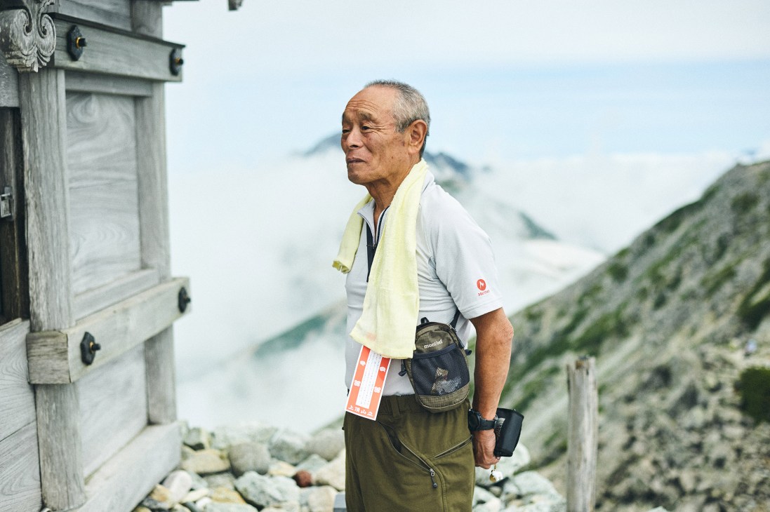 Older Japanese man in white shirt with yellow towel at mountain shrine overlooking Tateyama peaks.