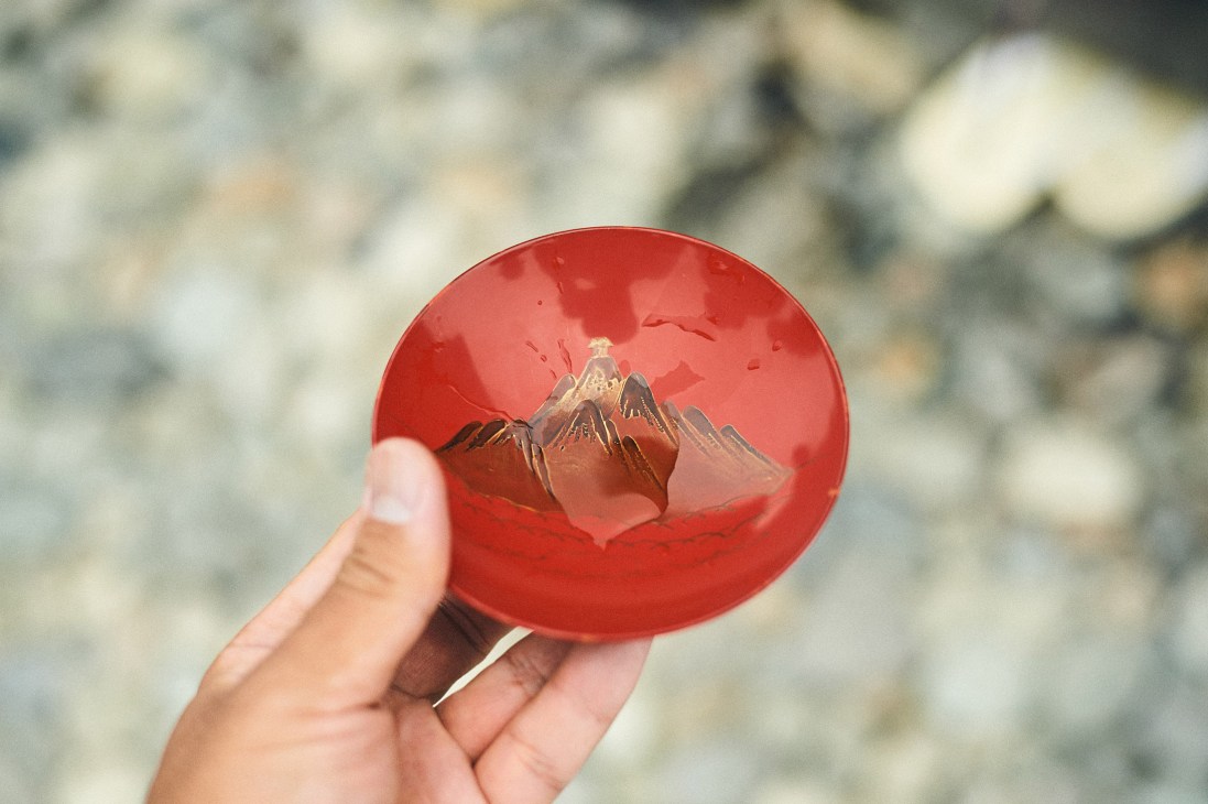 Hand holding a bright red lacquered sake cup with mountain landscape etched inside.