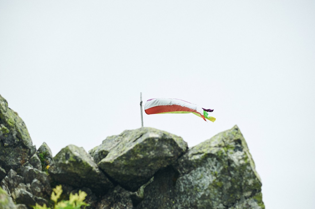 A colorful windsock flag flying above moss-covered rocks against a pale sky on a mountain peak.