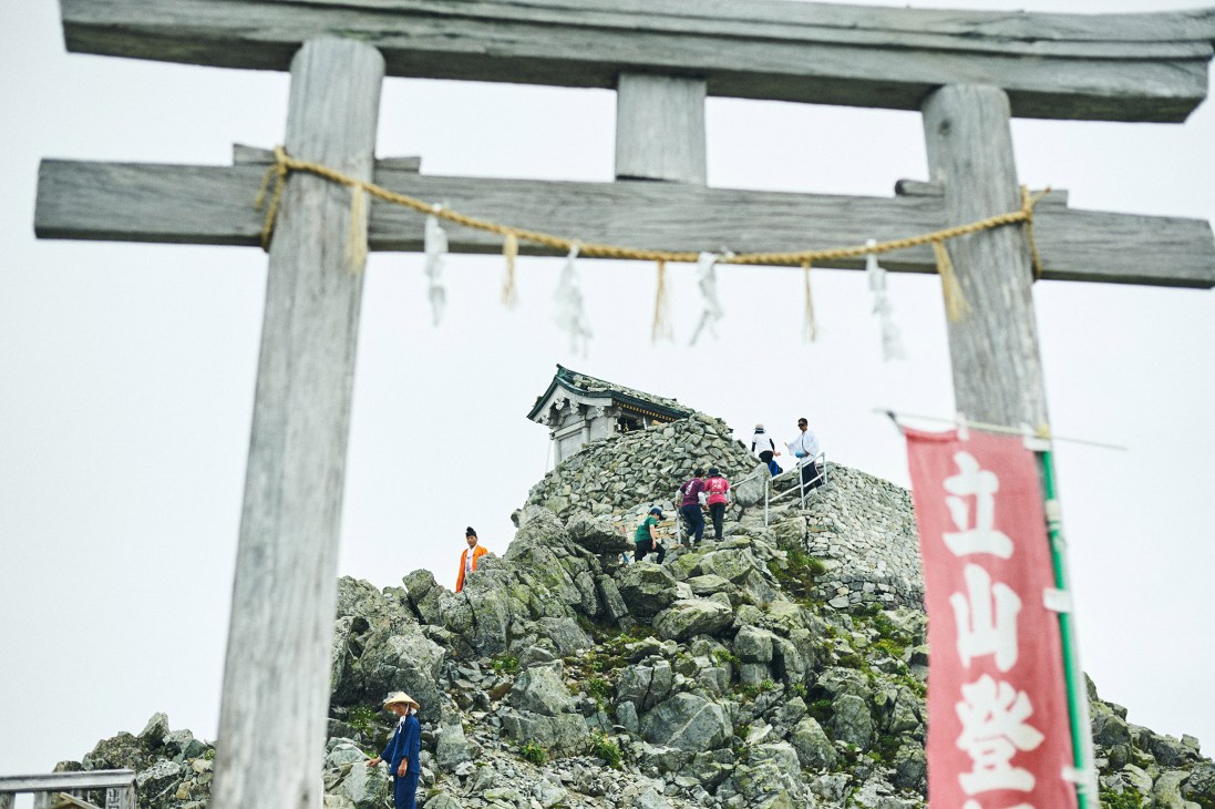 Hikers climbing rocky mountain peak with wooden torii gate and shrine structure visible against white sky.