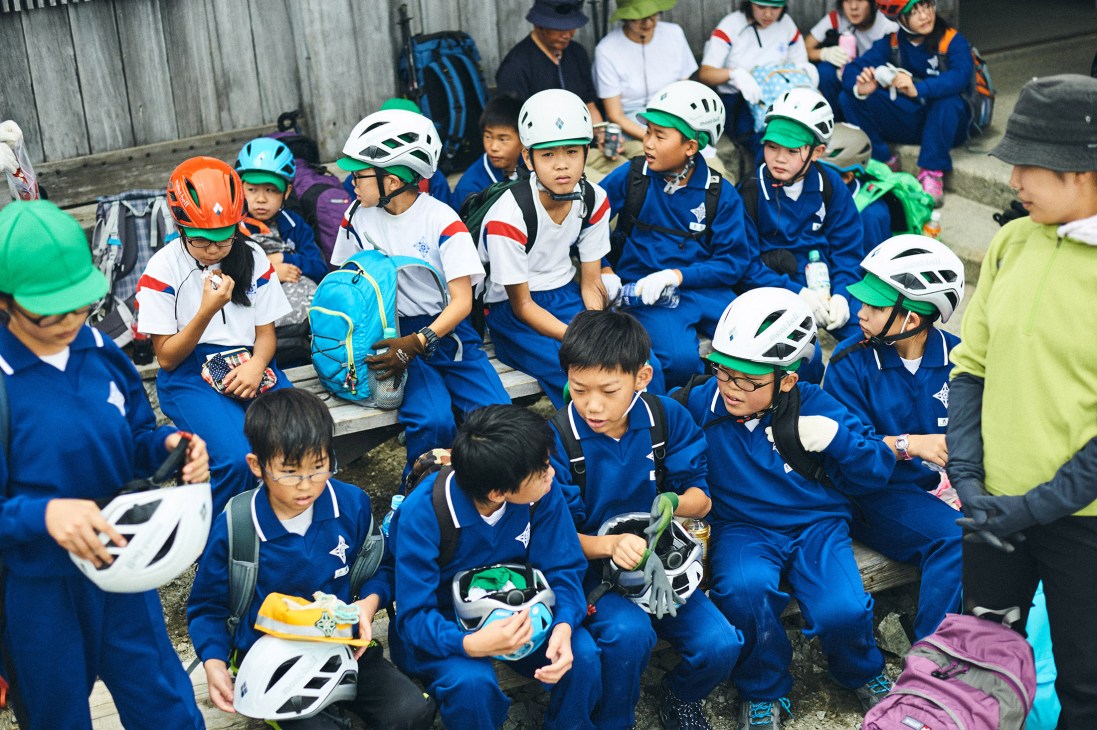 Schoolchildren wearing blue tracksuits and white safety helmets rest during a field trip on Tateyama mountain.