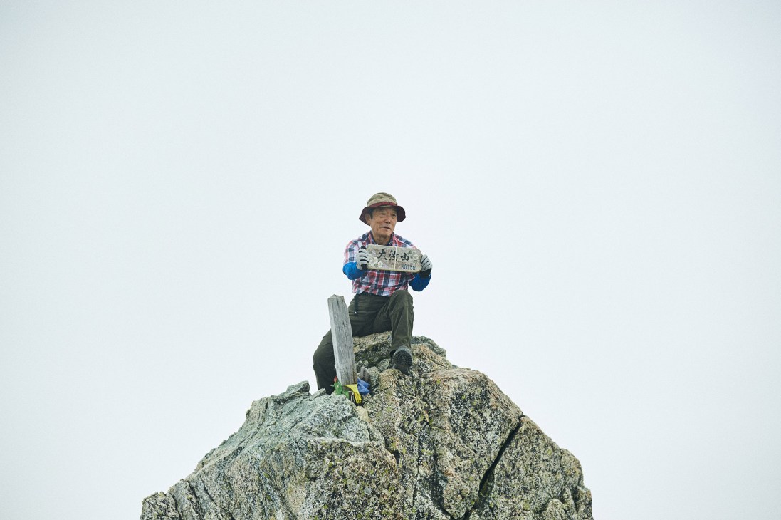 An older man in a plaid shirt and blue sleeves stands atop a rocky mountain peak at high altitude.