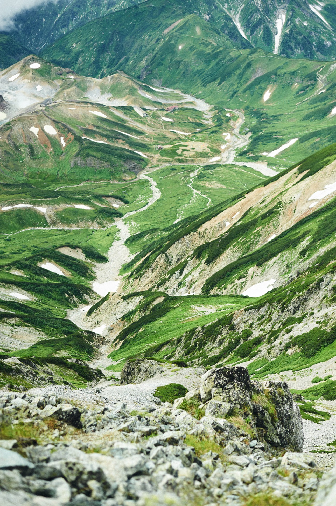 Aerial view of verdant alpine valleys with winding streams and patches of snow on the Tateyama mountain range.