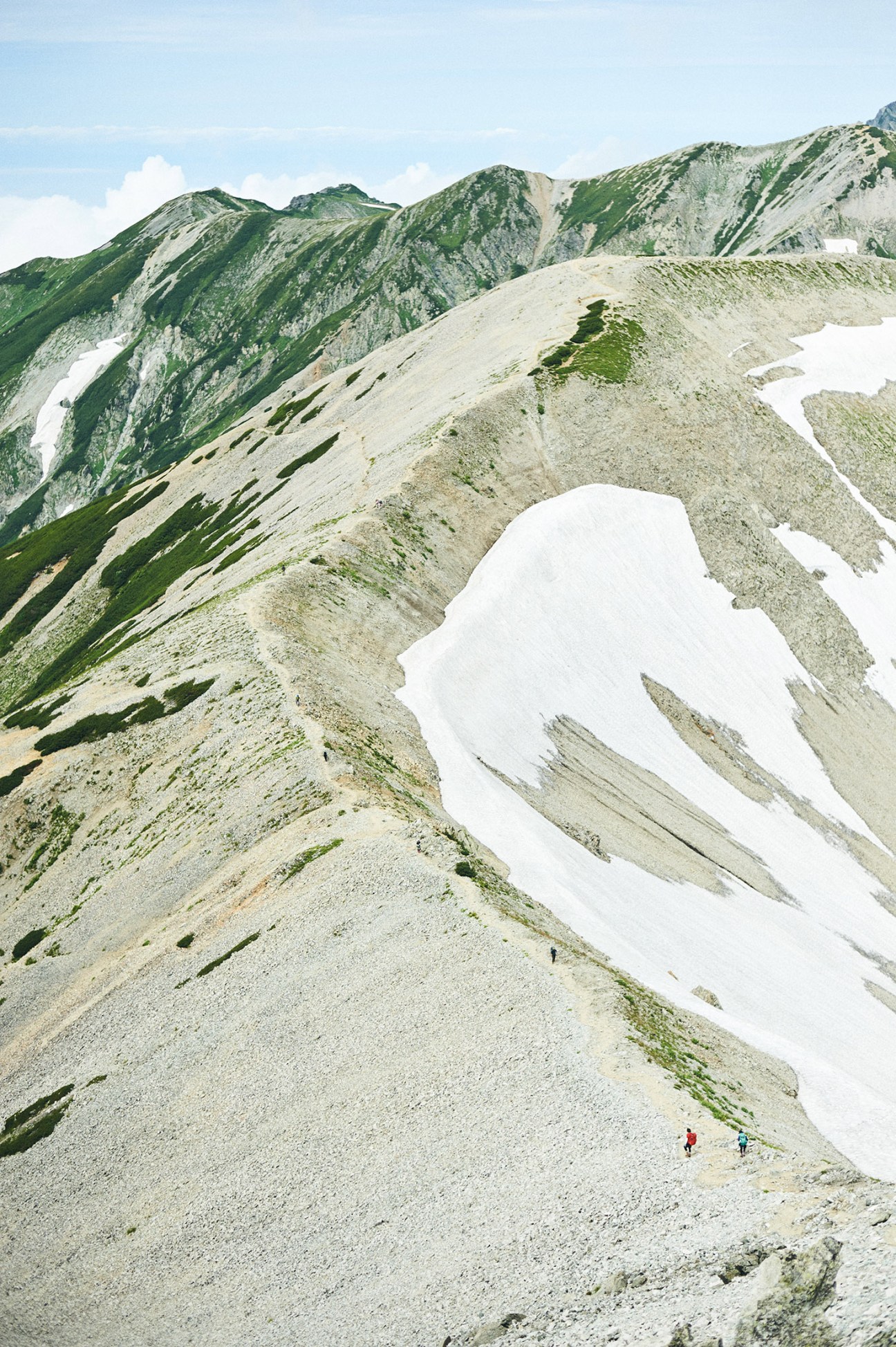 Hikers in colorful clothing traverse a rocky alpine slope with snow patches and verdant peaks in the background.