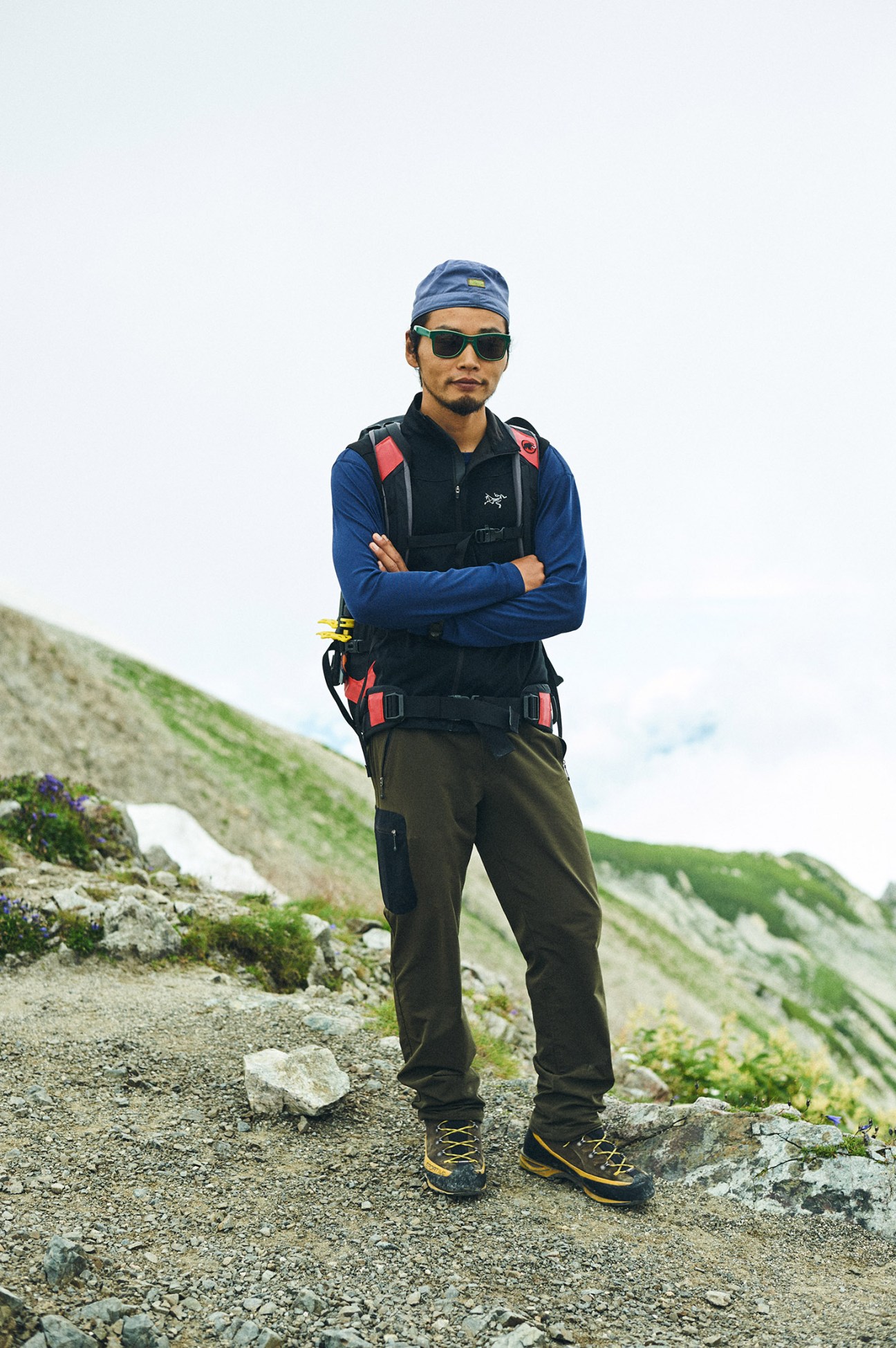 Guide Toshitaka Nakamura stands on a mountain trail wearing blue jacket, black vest, olive trousers and sunglasses.