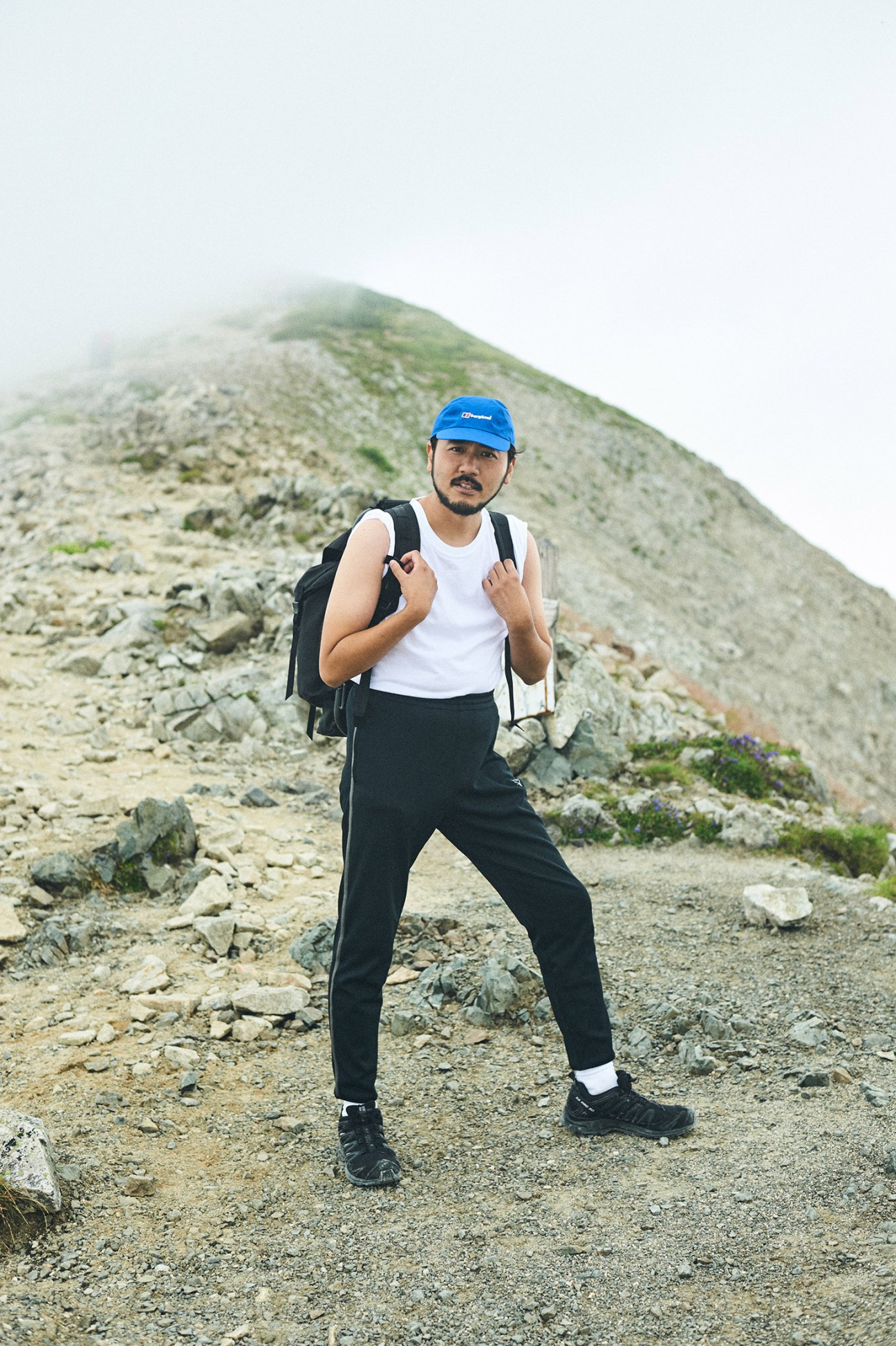 A hiker wearing a blue cap, white tank top, black pants and backpack stands on a rocky mountain trail with misty peaks in the background.