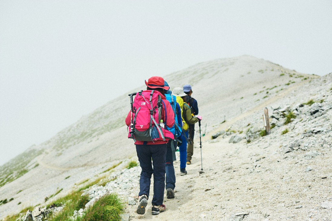 Three hikers in coordinated colored outfits walk single file up a rocky mountain trail on Tateyama.