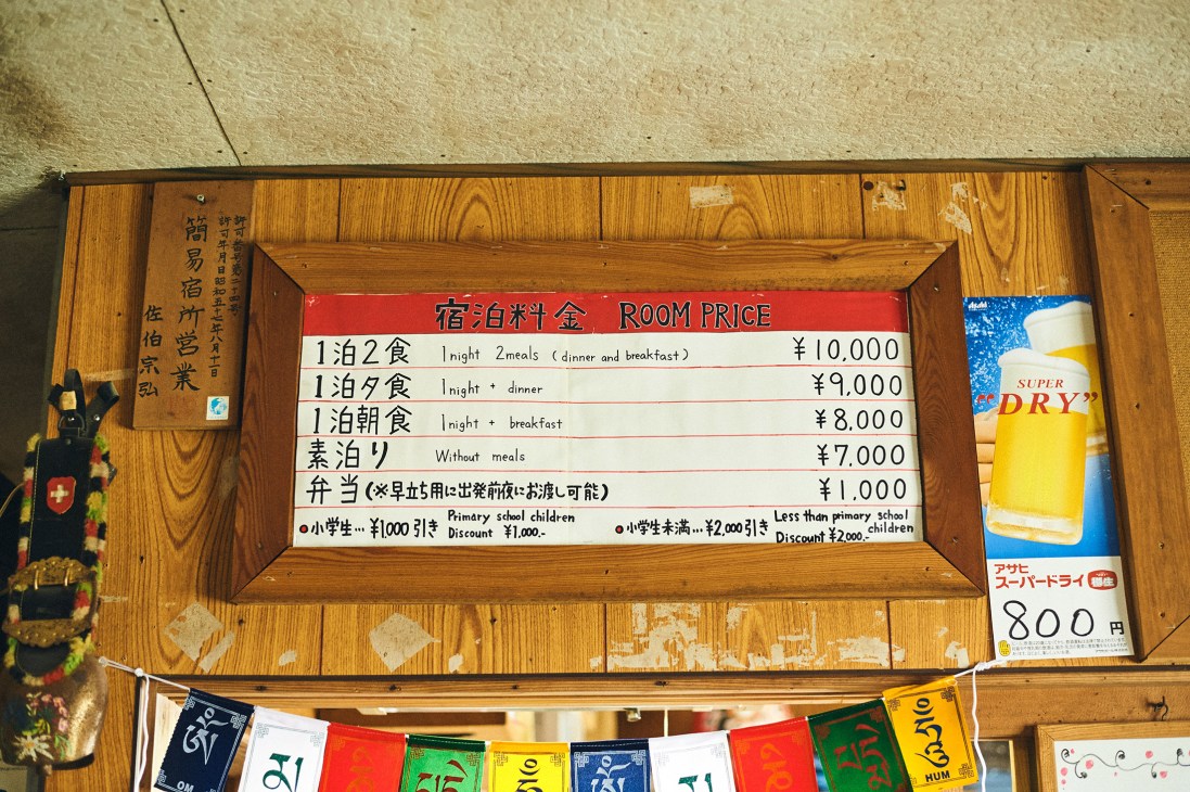 A wooden sign displaying room prices in Japanese and English for a mountain cottage.