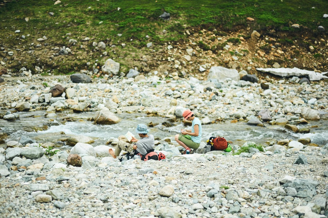 Two hikers rest on rocky ground beside a mountain stream in the Tateyama range.