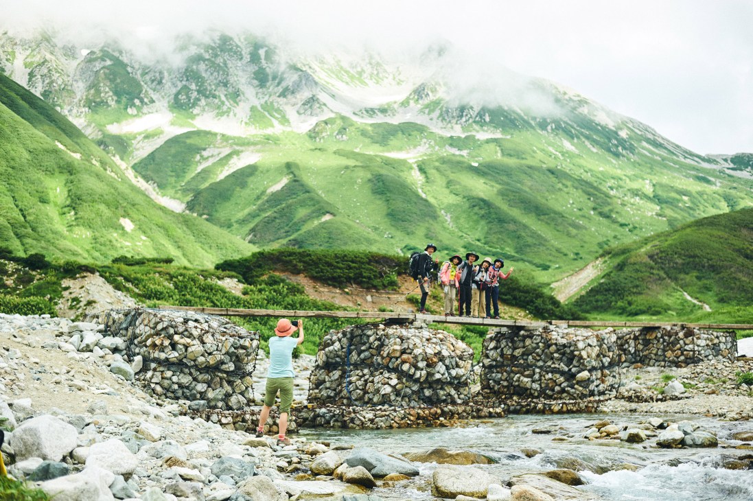 Hikers in colorful outdoor clothing stand on a wooden bridge over a mountain stream with verdant peaks in the background.