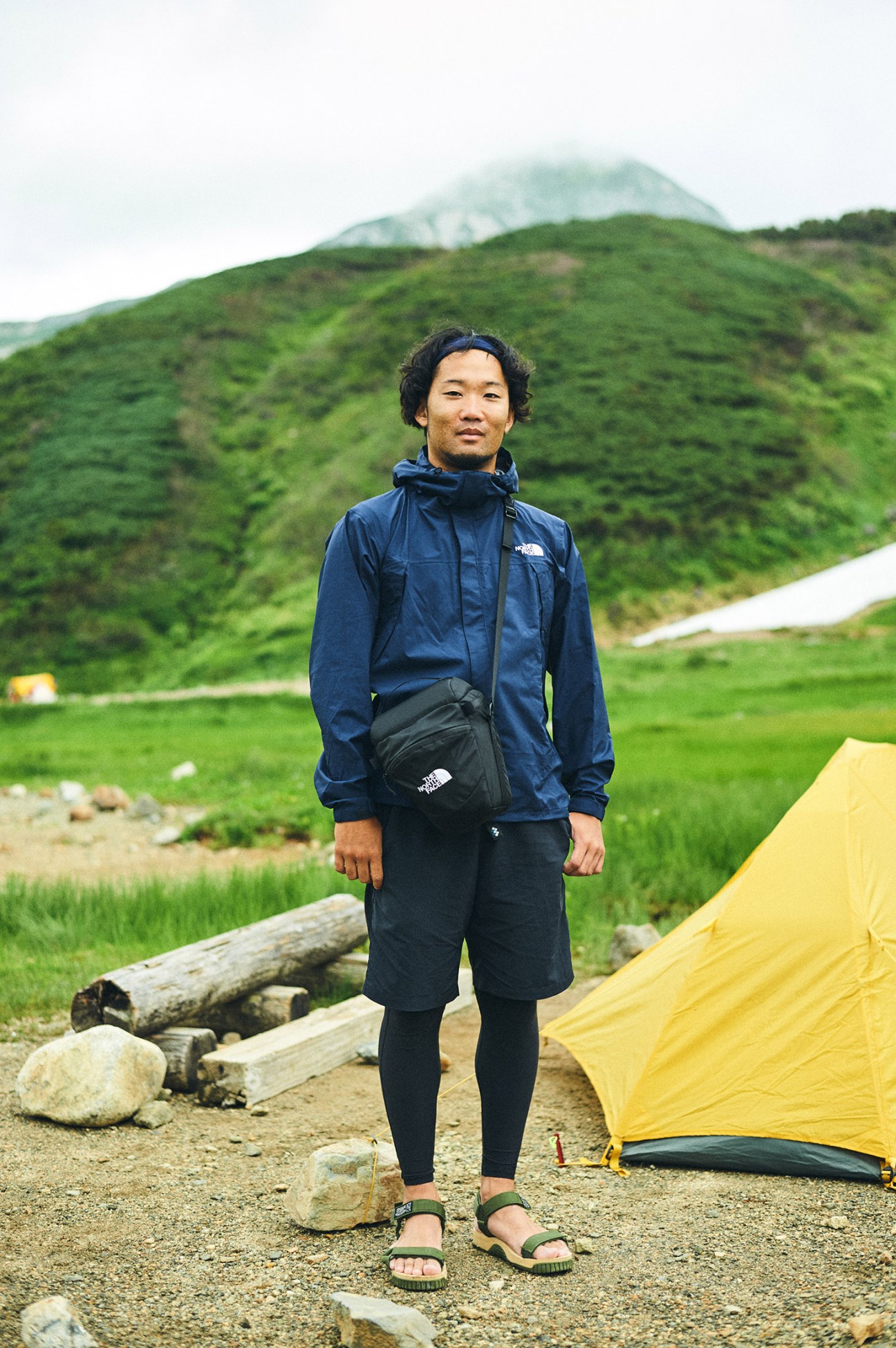 Man in blue North Face jacket standing at mountain camp with green slopes and tent behind him.