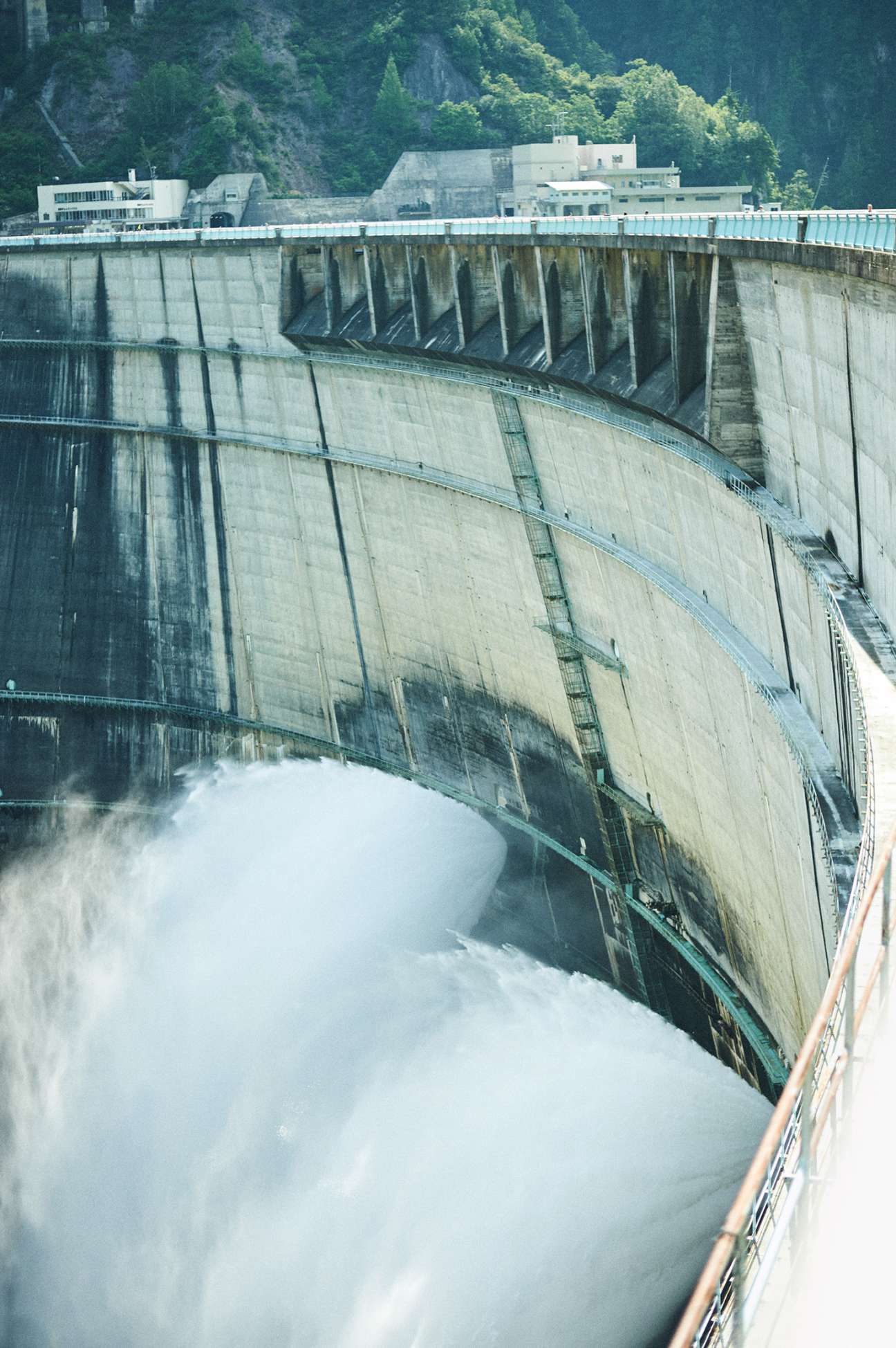Kurobe Dam's massive concrete face with water cascading from spillways, forested mountains rising behind.