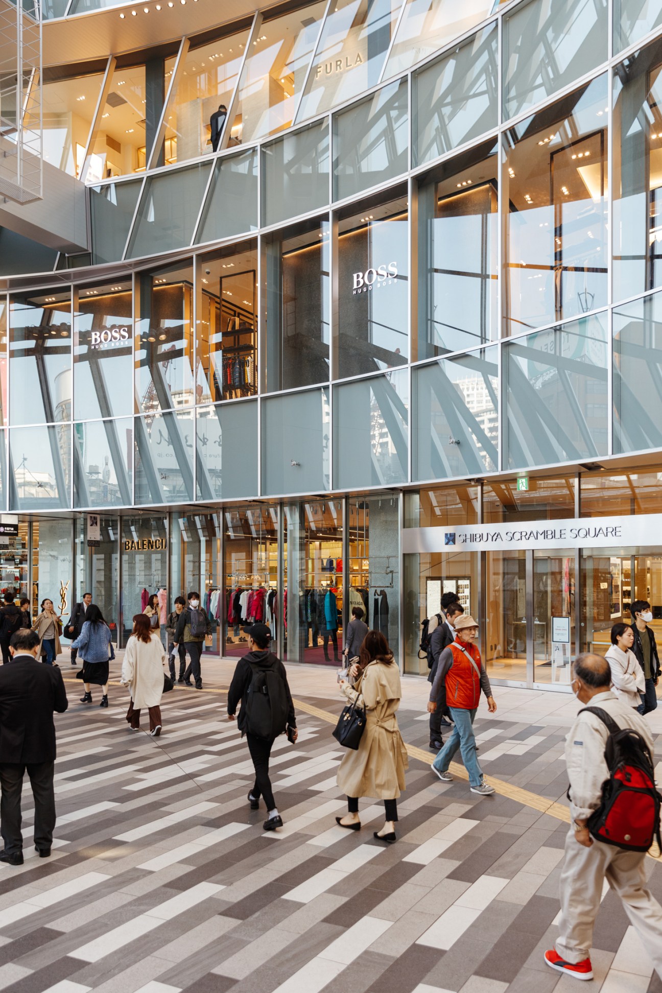 Modern multi-level shopping mall interior with glass facades, luxury brand stores including Boss and Balenciaga