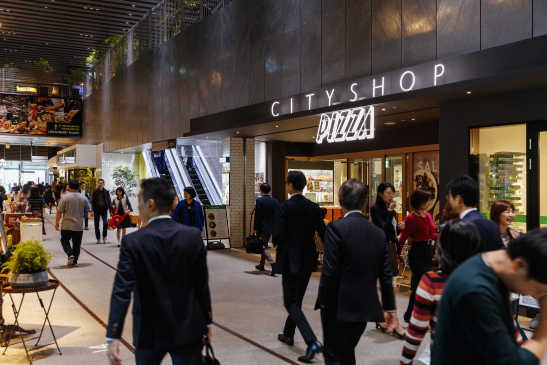 Busy shopping concourse at Japanese train station with City Shop Pizza storefront and commuters walking