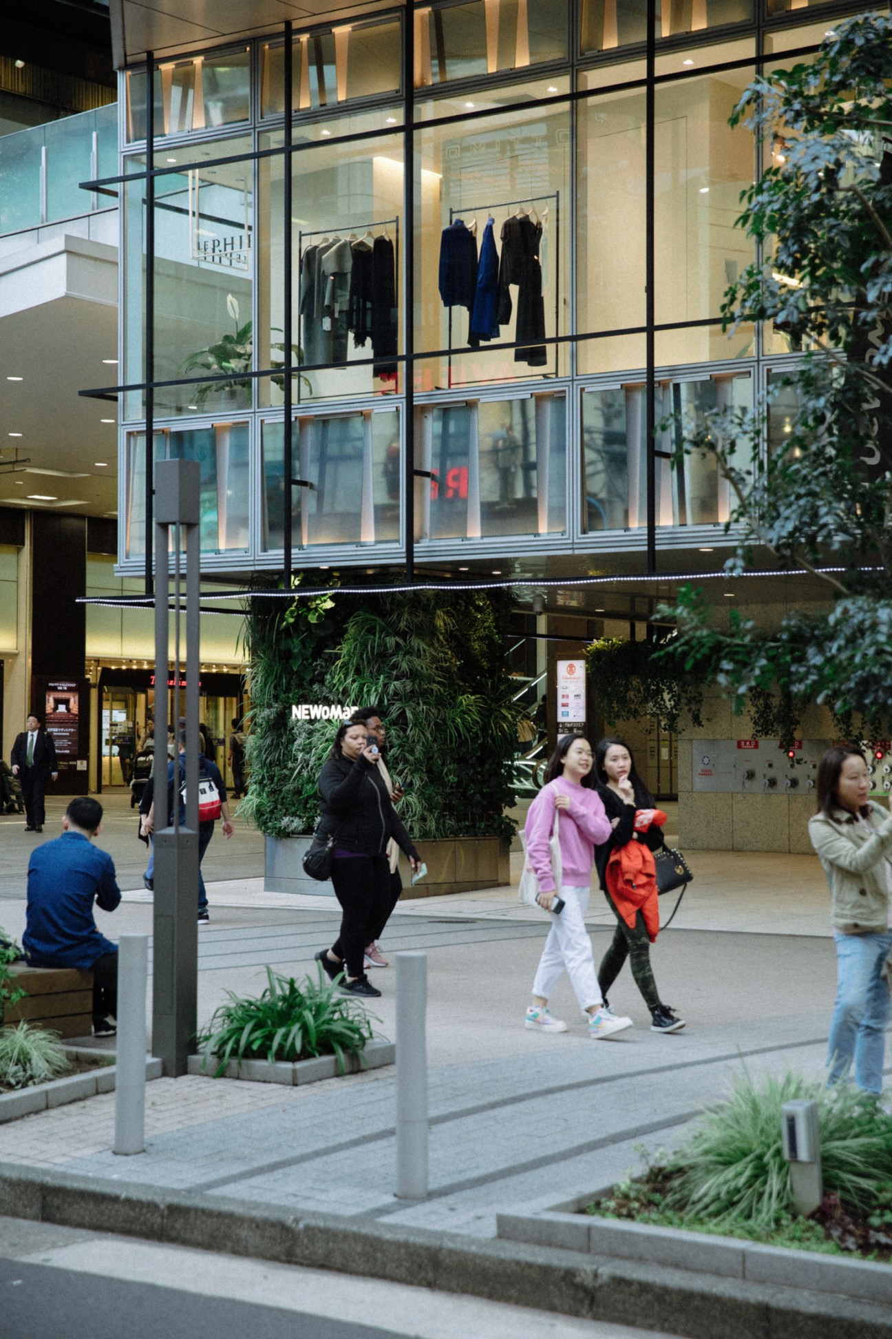 People walking through a modern shopping mall with glass storefronts and greenery