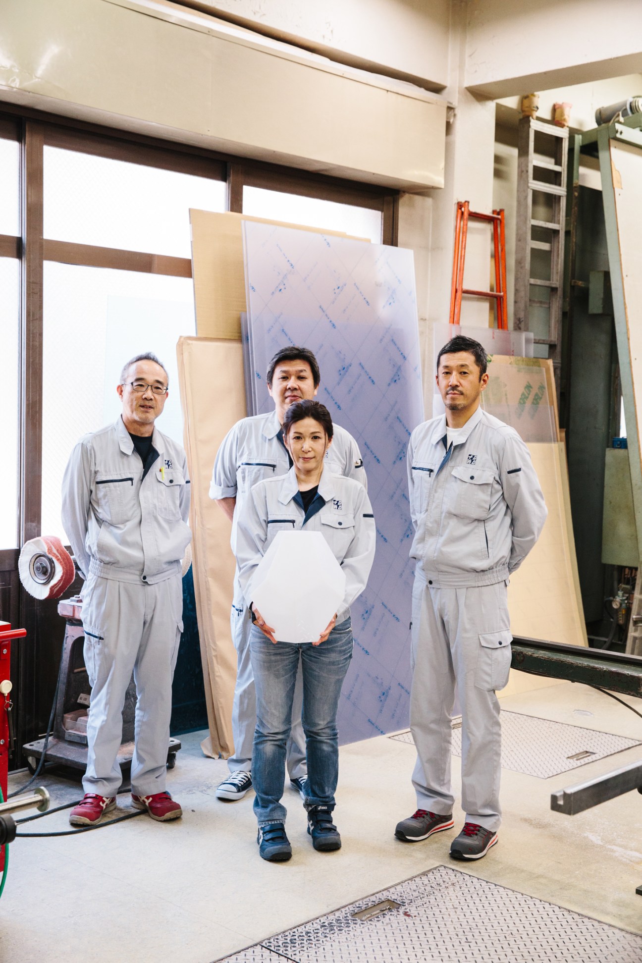 Four craftspeople in grey work uniforms stand in a workshop holding white papers, surrounded by wooden materials