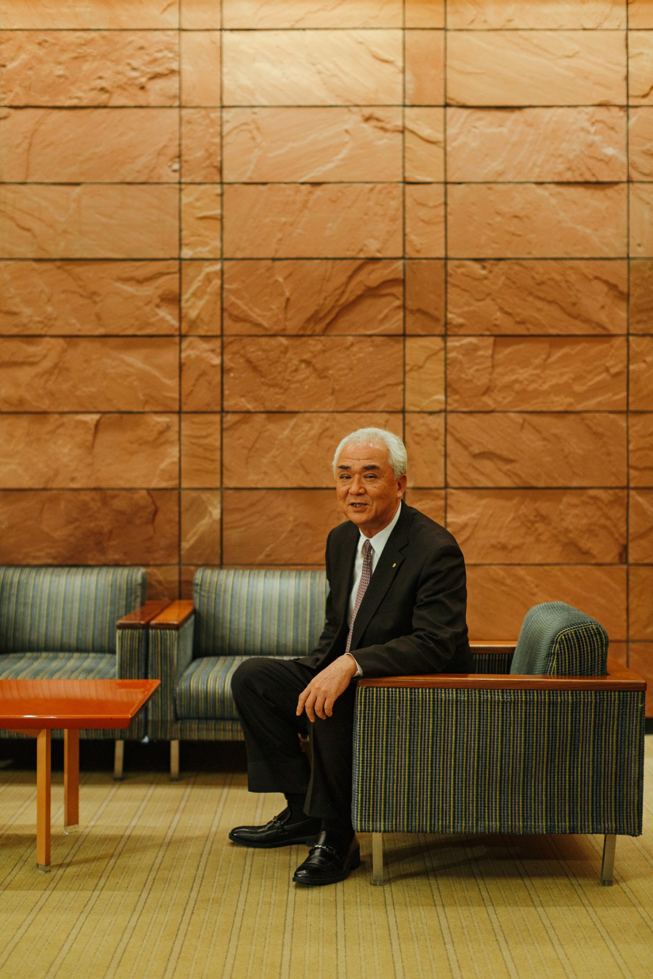 Elderly Japanese man in dark suit sitting on striped upholstered chair in modern lobby with wood-paneled walls