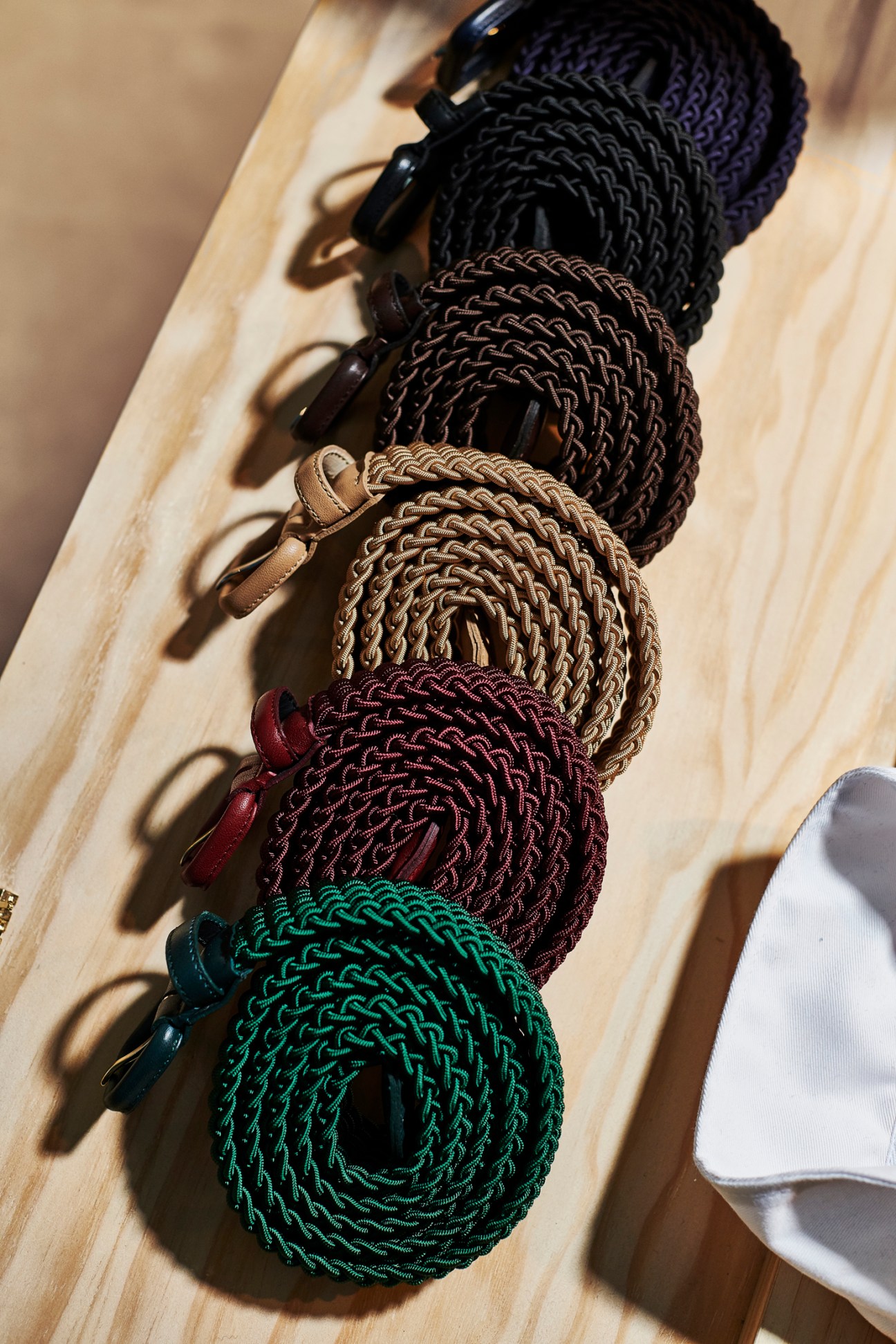 Five braided belts in different colors displayed on light wood, with metal buckles.