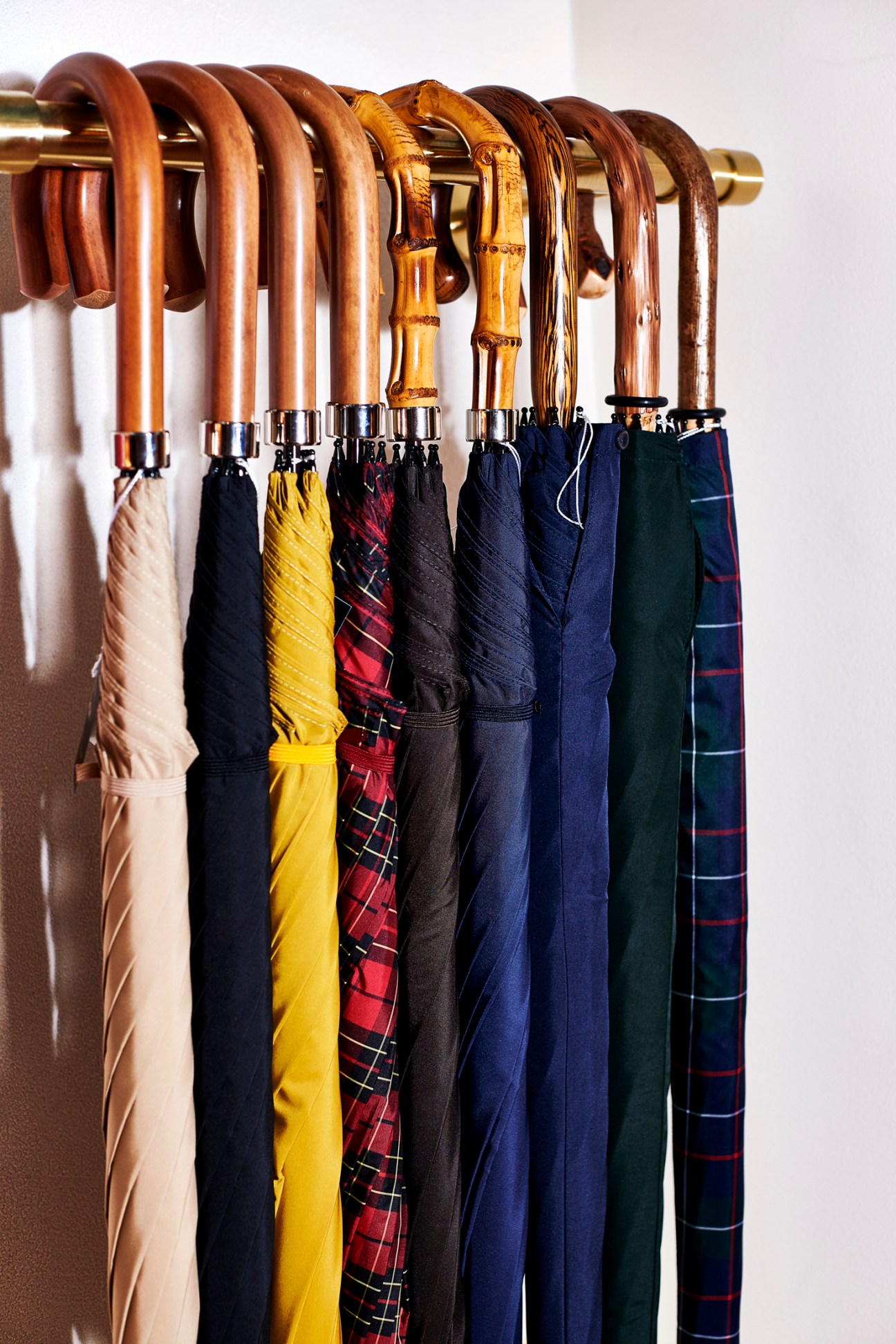 Row of Fox umbrellas with wooden handles in various colors including cream, black, yellow, red tartan, and navy displayed on a brass rod.