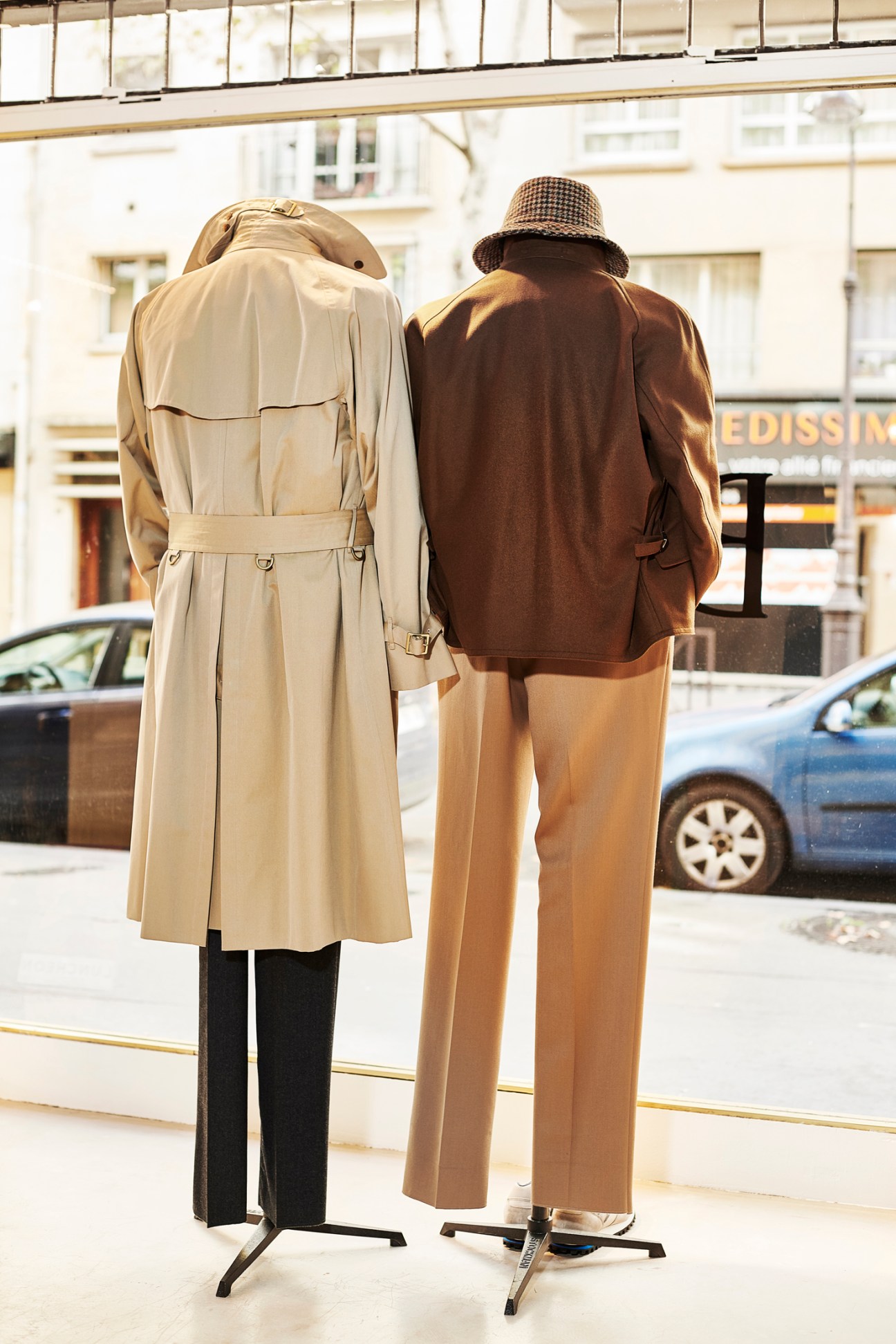 Two dressed mannequins in a storefront window displaying menswear including a trench coat and blazer.