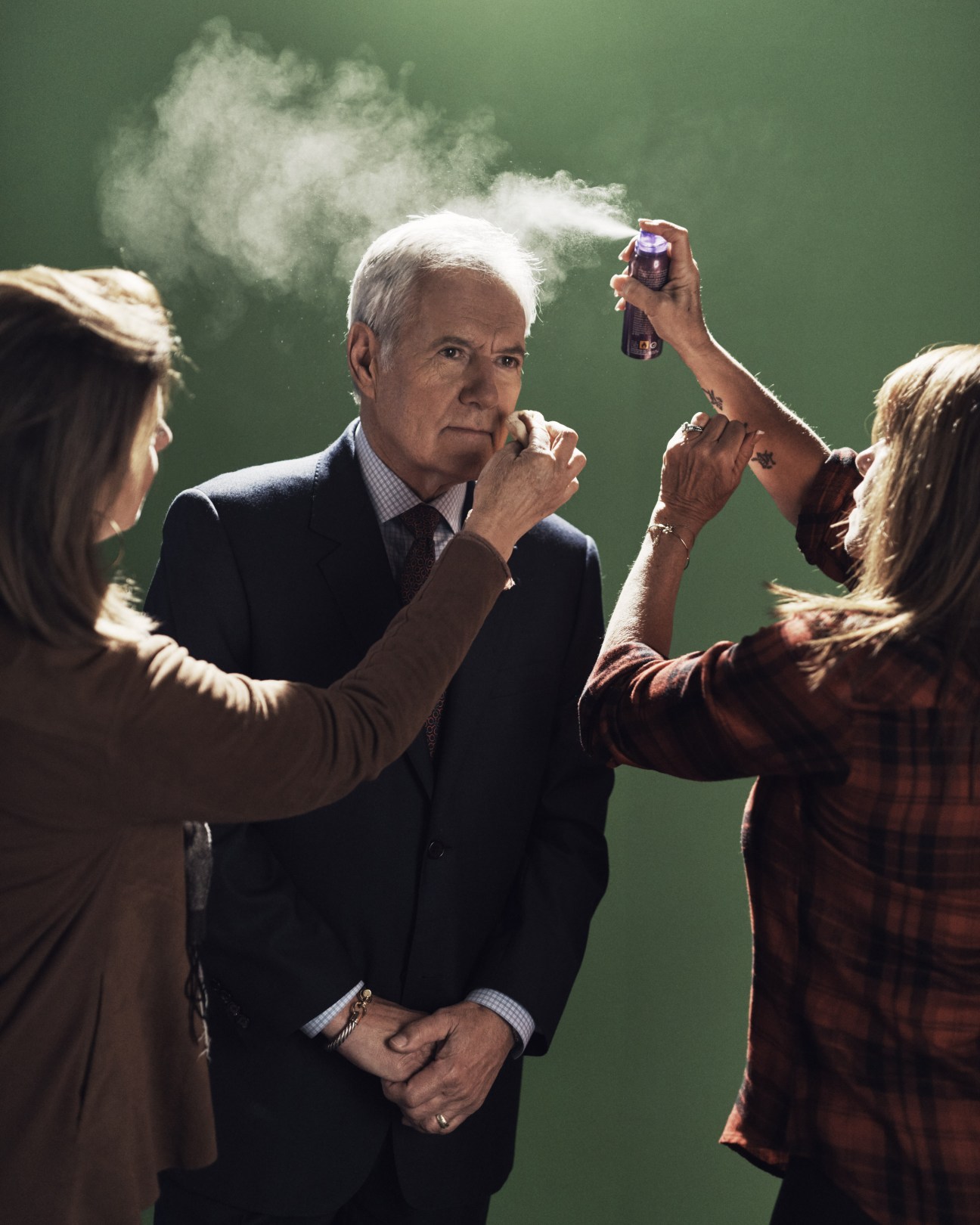 Jeopardy! host Alex Trebek being prepared for taping by two women applying makeup and hair spray against a green backdrop.