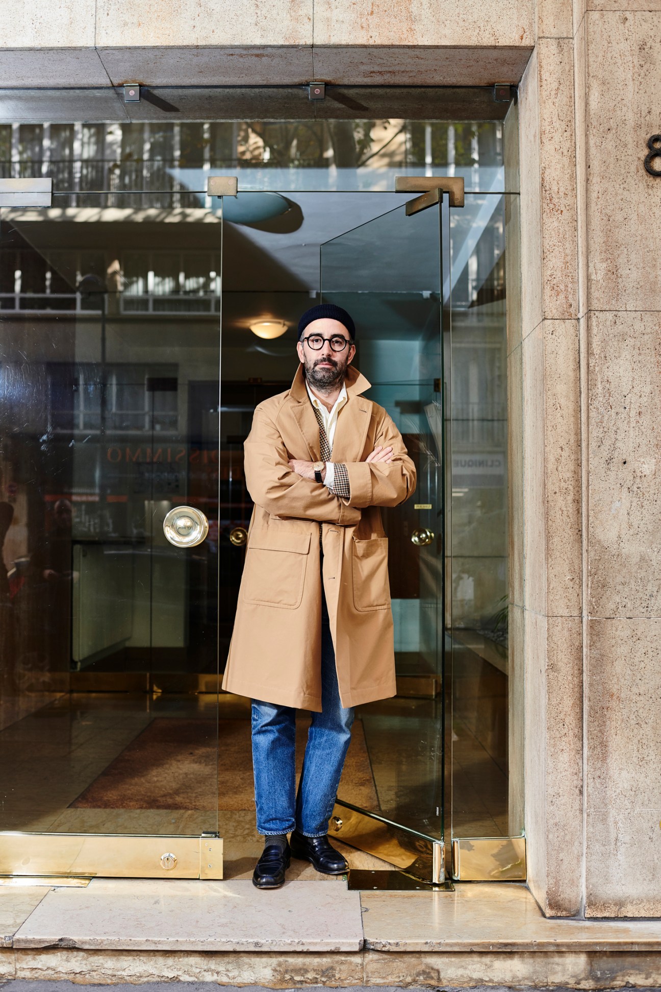 Man with glasses wearing a camel coat and jeans stands in front of Beige Habilleur store entrance in Paris.