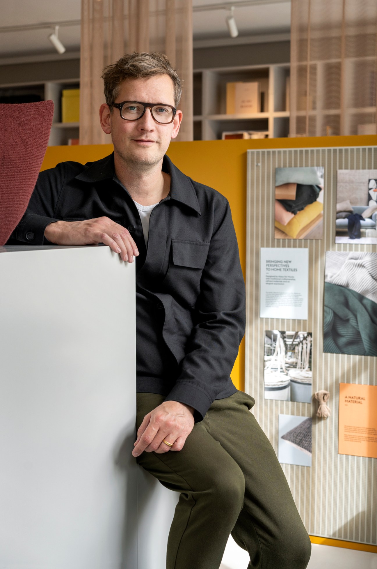 Man in black shirt and glasses sitting on white furniture in modern office with design displays and bookshelves
