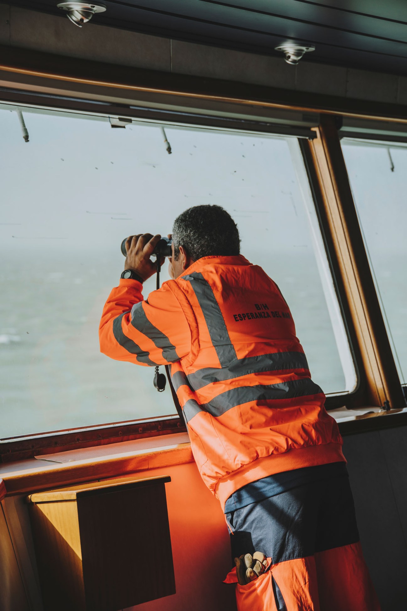 Crew member in orange safety gear using binoculars to scan the horizon from the bridge of the Esperanza del Mar.