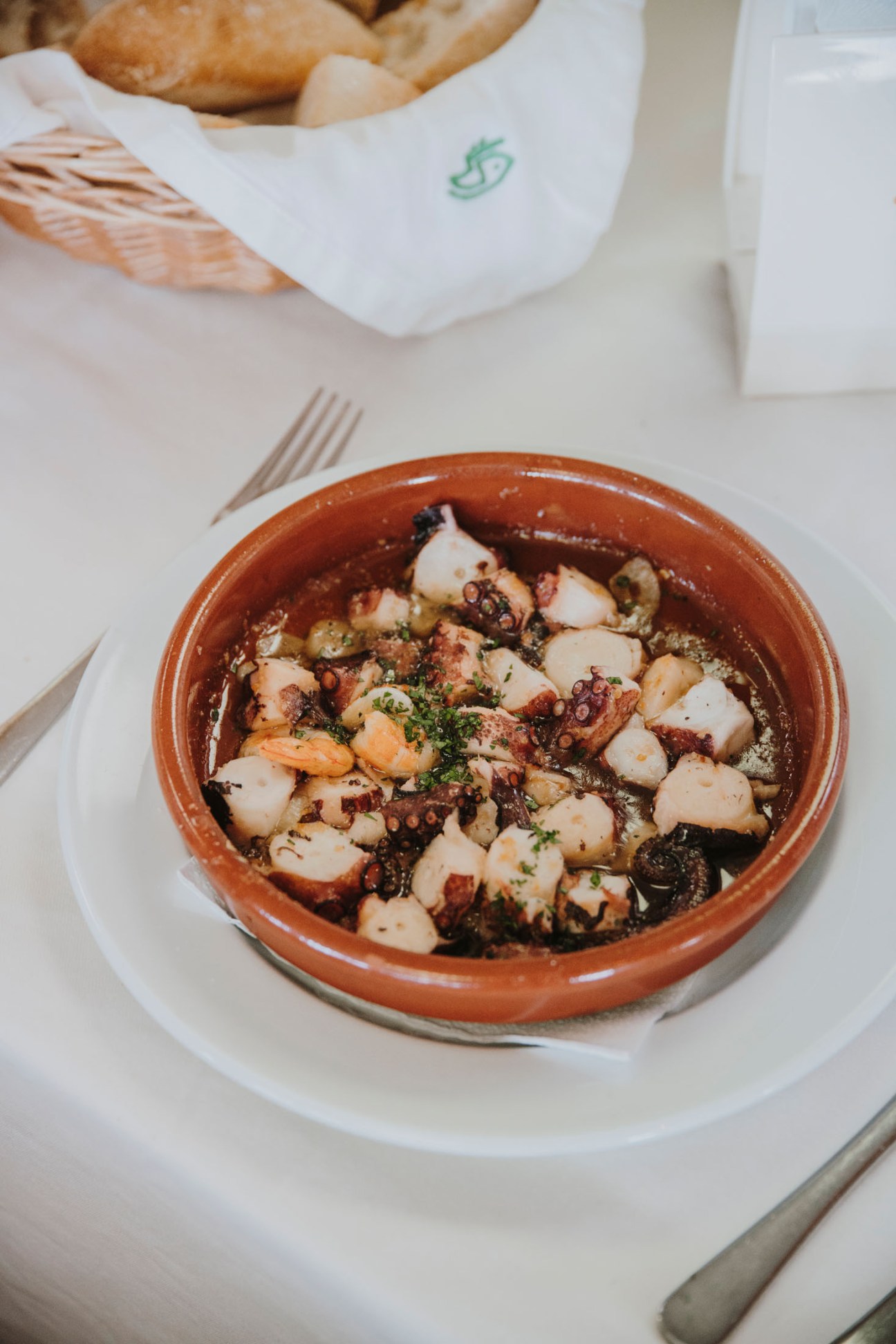 Octopus dish with tentacles and vegetables in a terracotta bowl on white plate with bread basket in background.