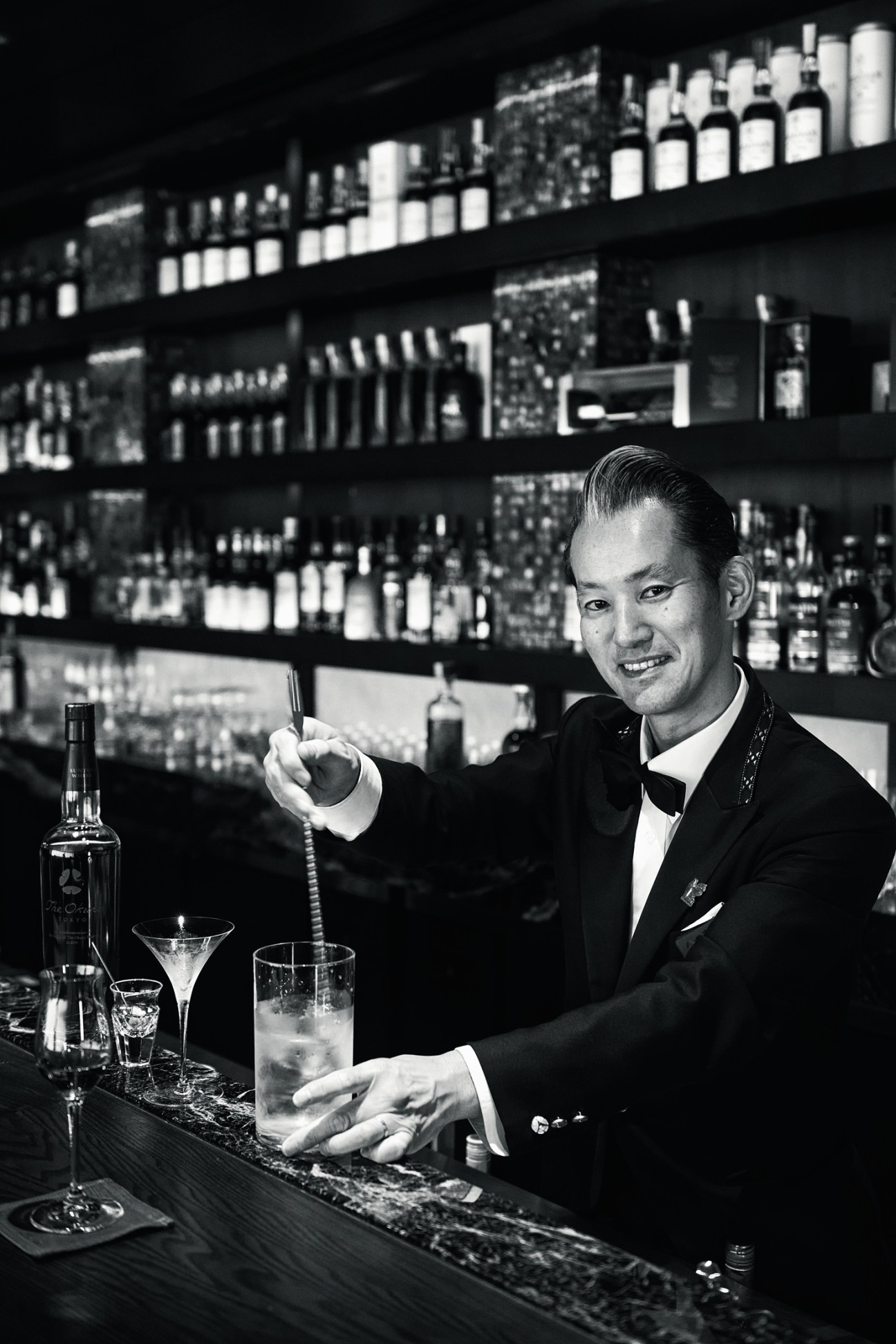 Portrait of Masakazu Takachi, a veteran bartender at The Okura Tokyo, mixing drinks behind a well-stocked bar