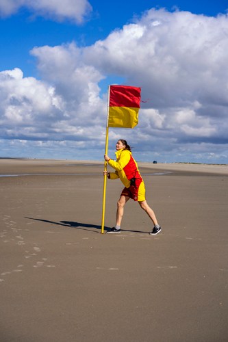 Lifeguard in red and yellow uniform holding rescue flag on windy beach