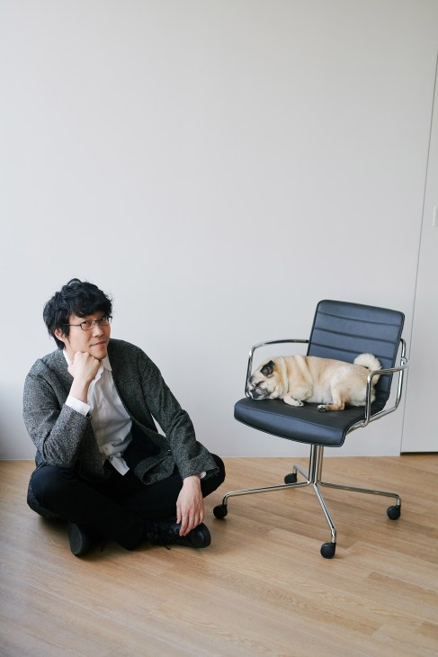 Oki Sato crouches beside a modern office chair on which his pug-chihuahua mix dog Kinako sits, against a minimalist white background.