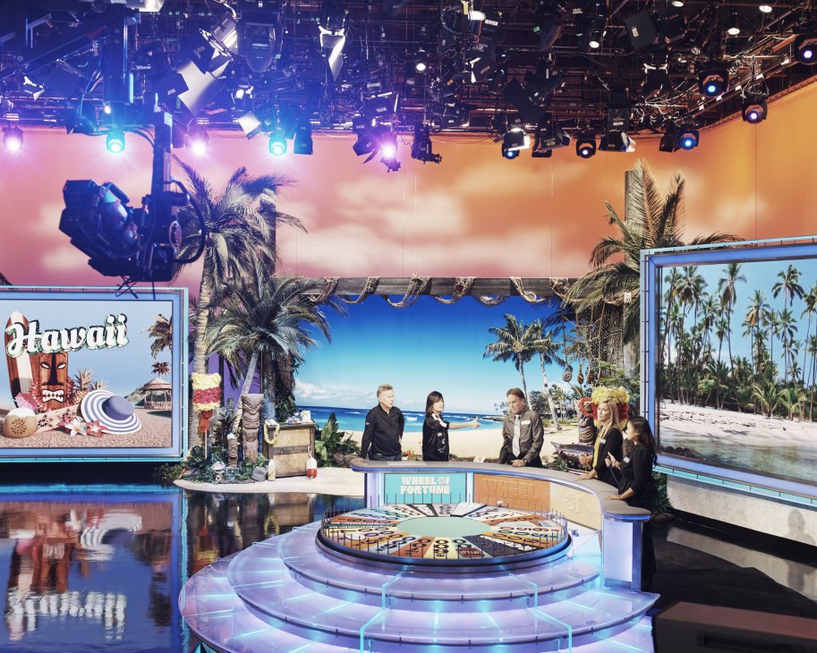 Behind-the-scenes rehearsal on the Wheel of Fortune set with Hawaii-themed backdrop, professional lighting rigs, and crew members preparing the stage.