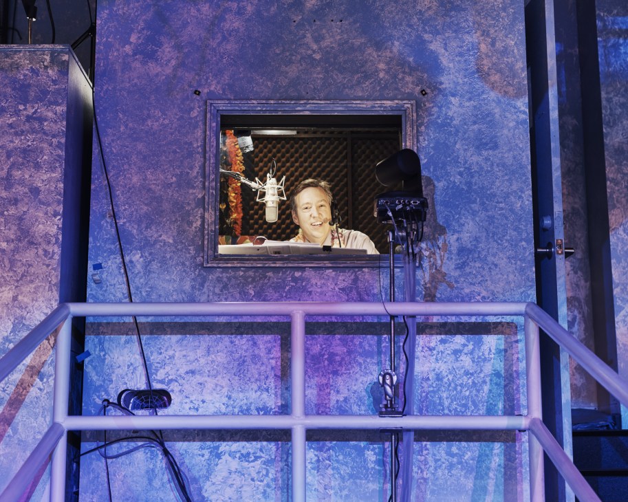 Wheel of Fortune announcer Jim Thornton speaking into a microphone in a purple-lit booth