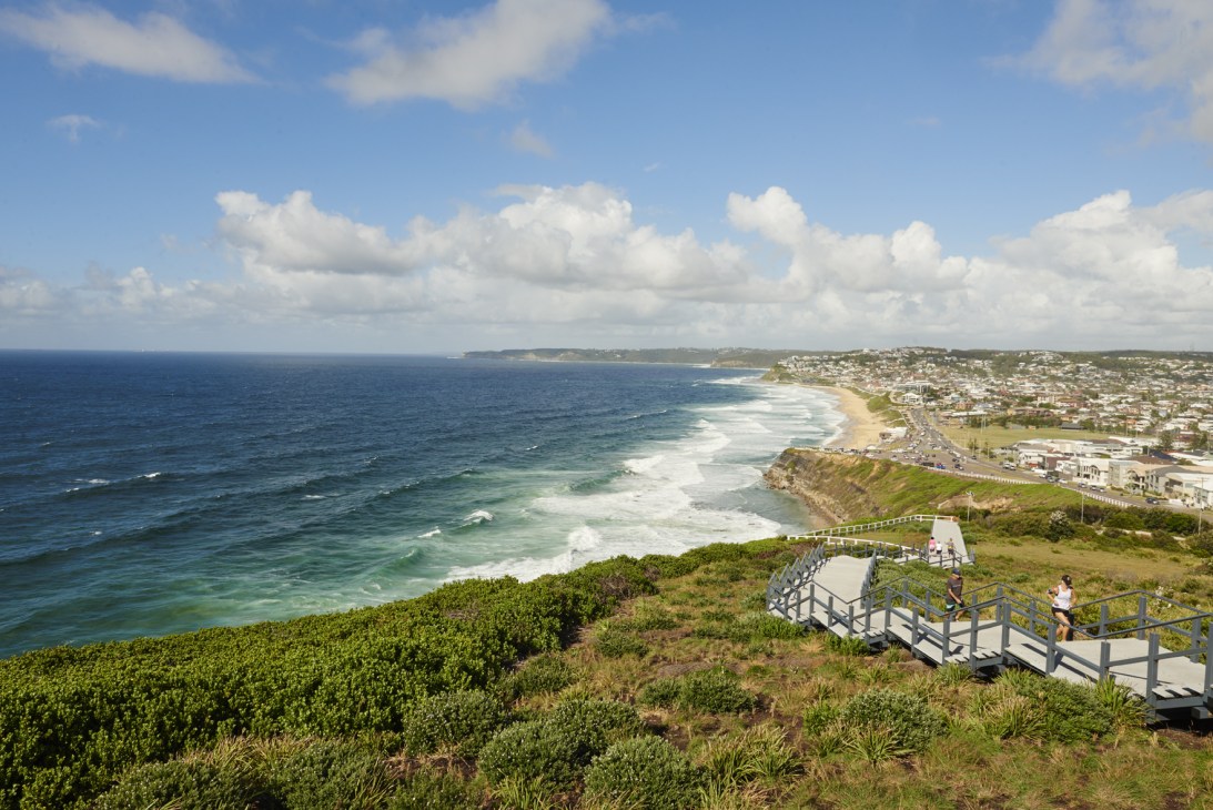 Coastal cliffs with wooden walkway and stairs overlooking beach and ocean waves under blue sky with clouds.
