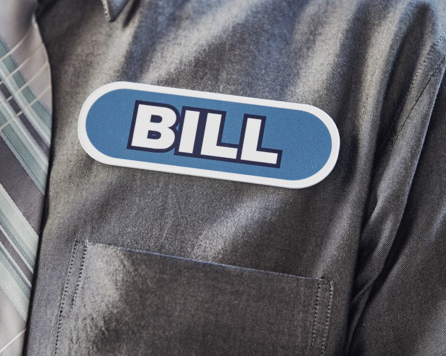 A person wearing a gray jacket with a blue "BILL" name badge celebrates with arms raised in triumph.