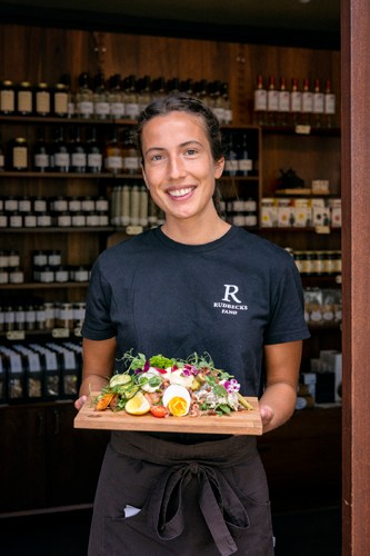 Smiling woman in dark shirt holding wooden board with colorful salad in front of shelved products