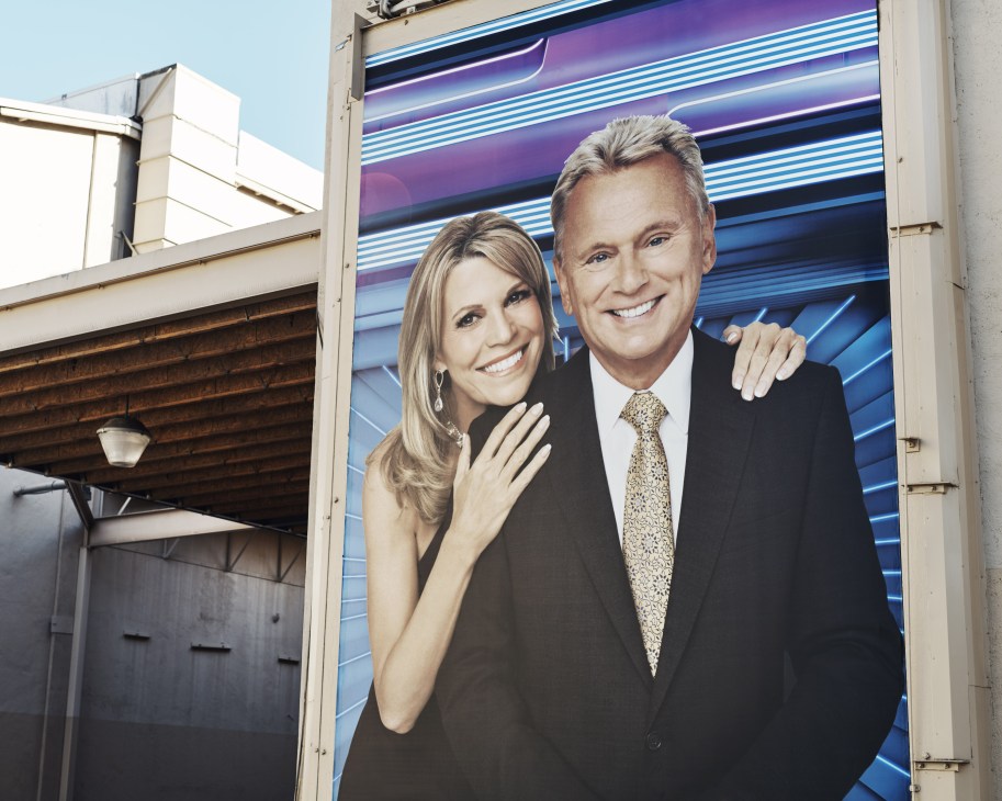Pat Sajak and Vanna White, hosts of Wheel of Fortune, smiling together on set with purple and blue striped backdrop