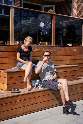 Two people enjoying drinks on wooden steps outside a modern building with glass railings